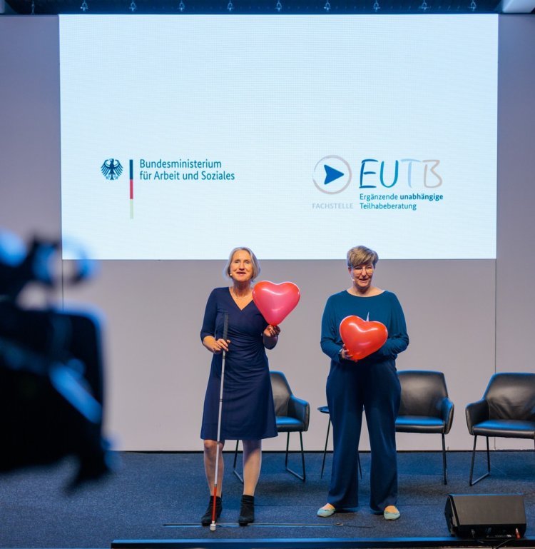 zwei Frauen sind mit Herz-Luftballons auf einer Bühne. Im Hintergrund ist das BMAS und das EUTB-Logo zu sehen